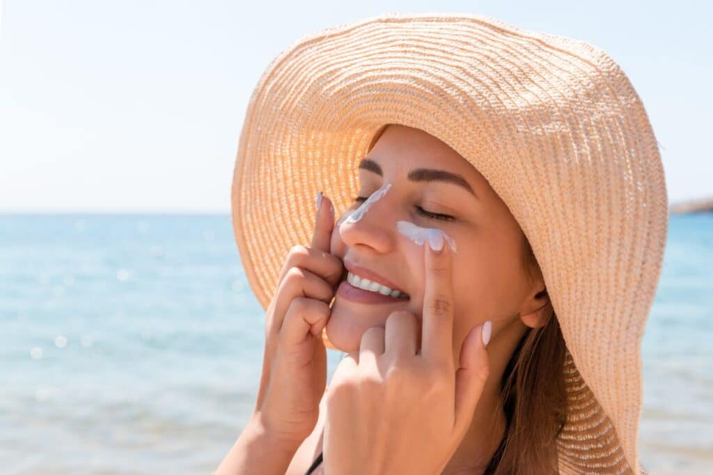 une femme souriante avec un chapeau applique de la crème solaire sur son visage